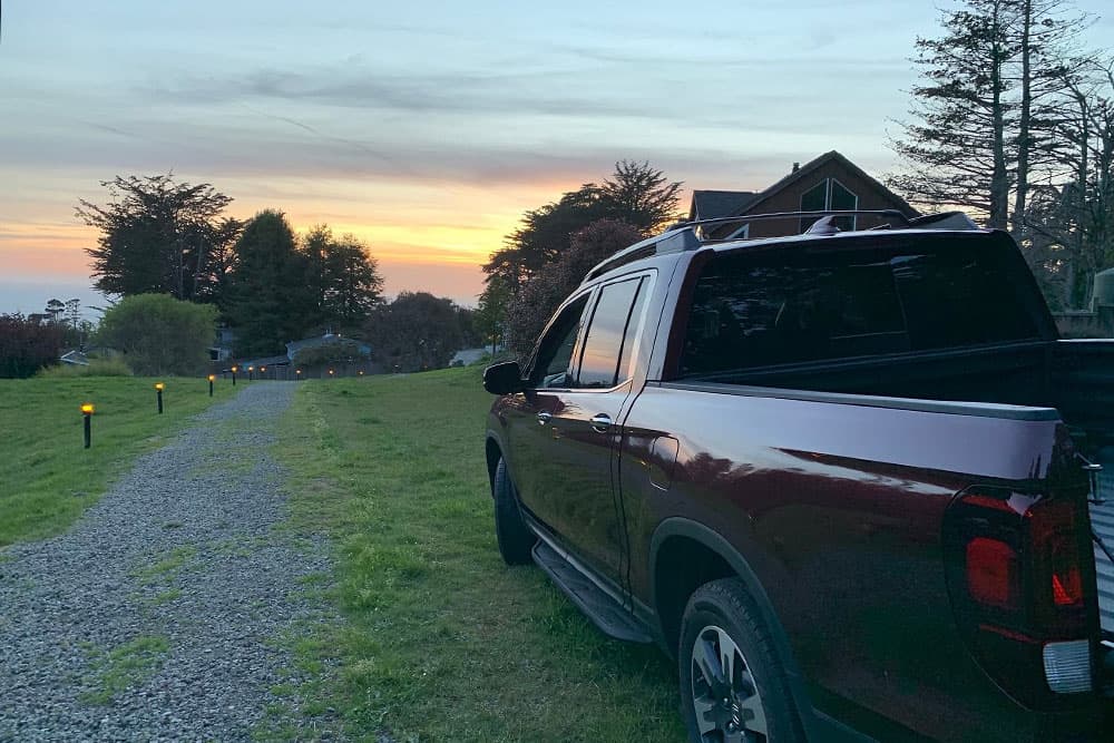 A maroon pickup truck parked on a gravel path at sunset, surrounded by trees and a house in the background.