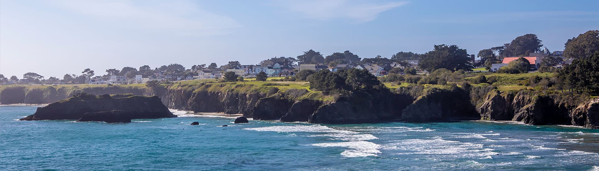 Scenic coastal view of Mendocino cliffs with waves crashing below and houses dotting the hillside above.