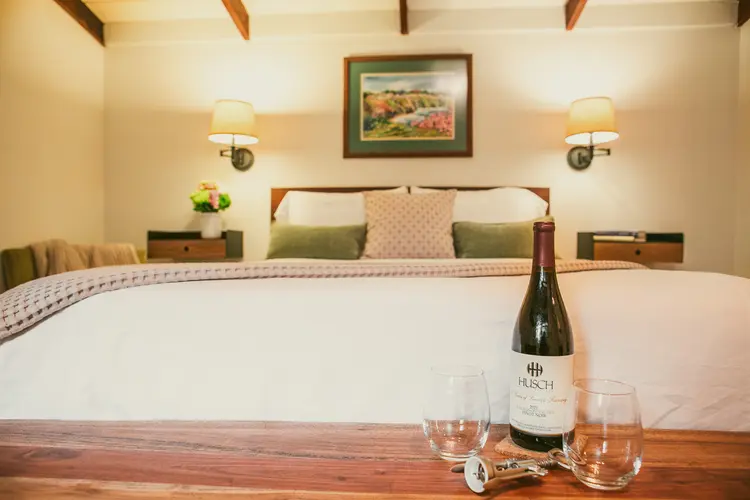 A cozy bedroom featuring a neatly made bed, a bottle of wine with glasses, and artwork on the wall.