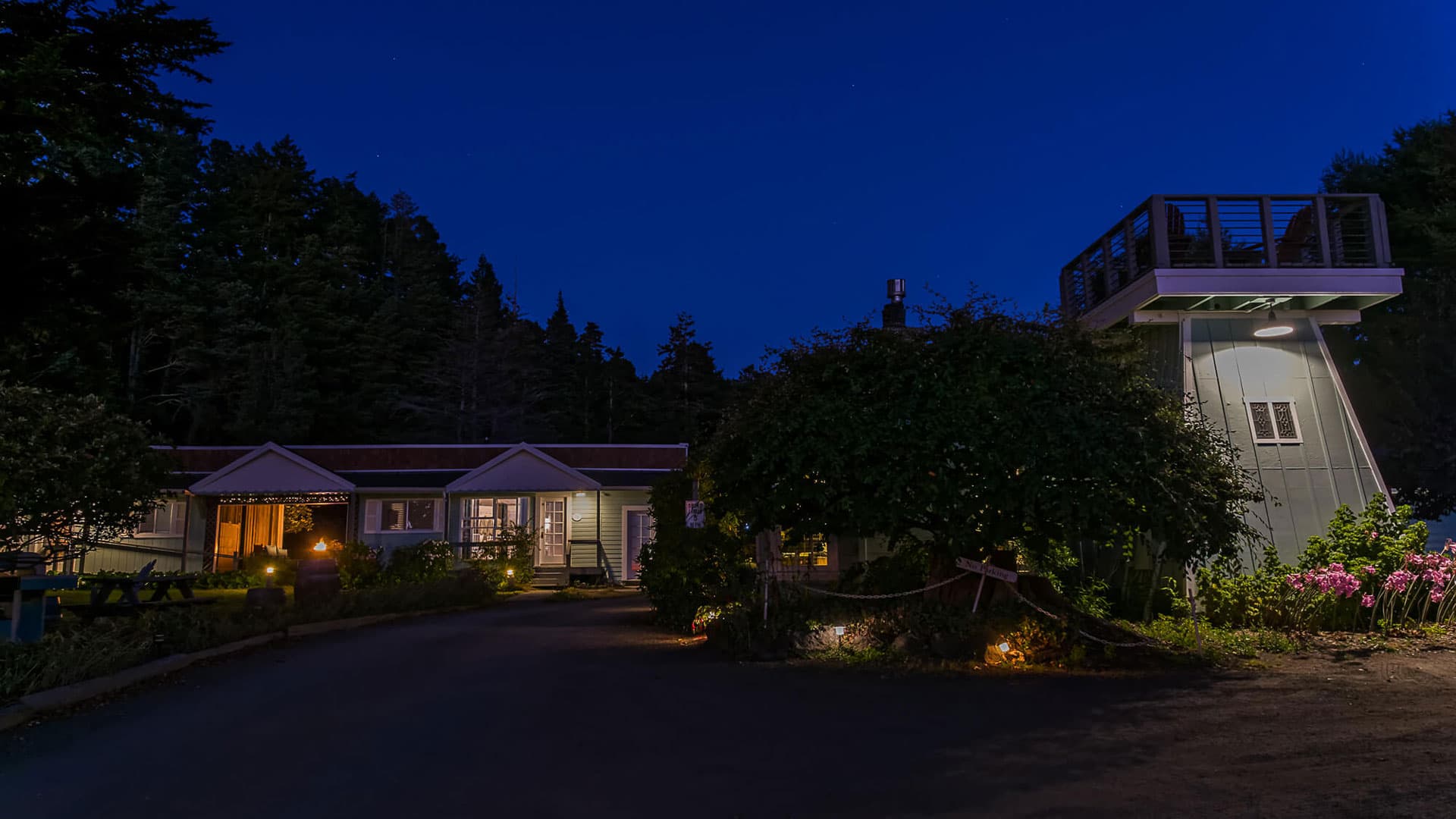Night view of a cozy lodge with soft exterior lighting, surrounded by trees, and a rooftop deck visible on the right.